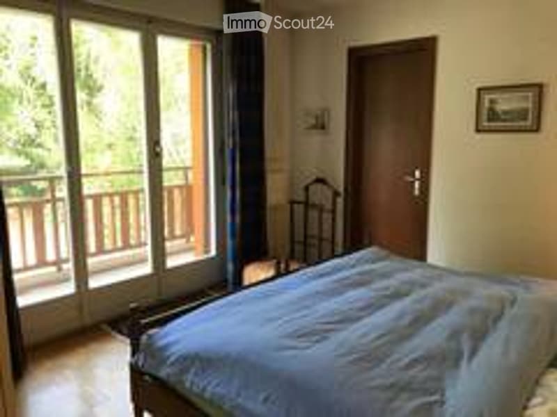 Cozy bedroom with a large bed, a wooden chair, and a picture frame on the wall. A sliding glass door opens to a balcony with wooden railings.