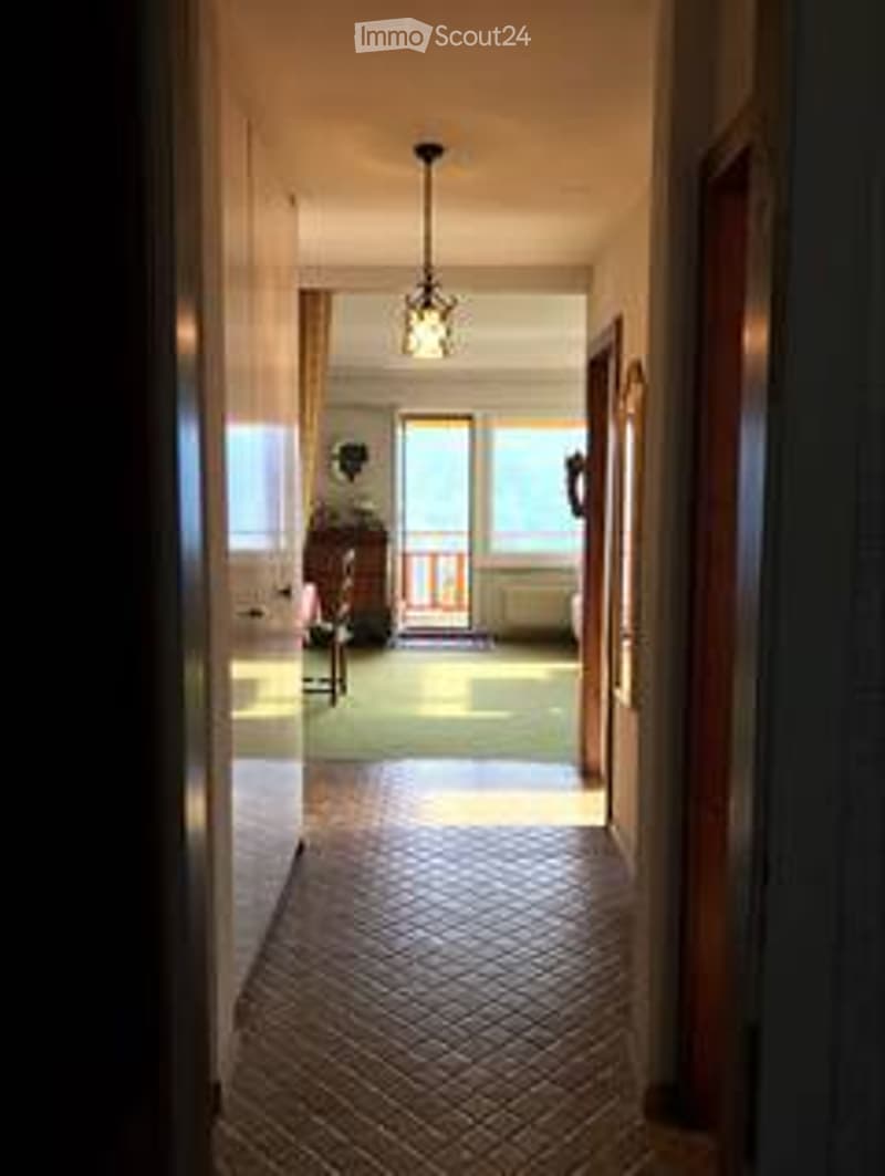 A long, well-lit hallway with a patterned tile floor, wooden doors on both sides, and a chandelier hanging from the ceiling. The hallway leads to a room with a visible sofa and a small table.