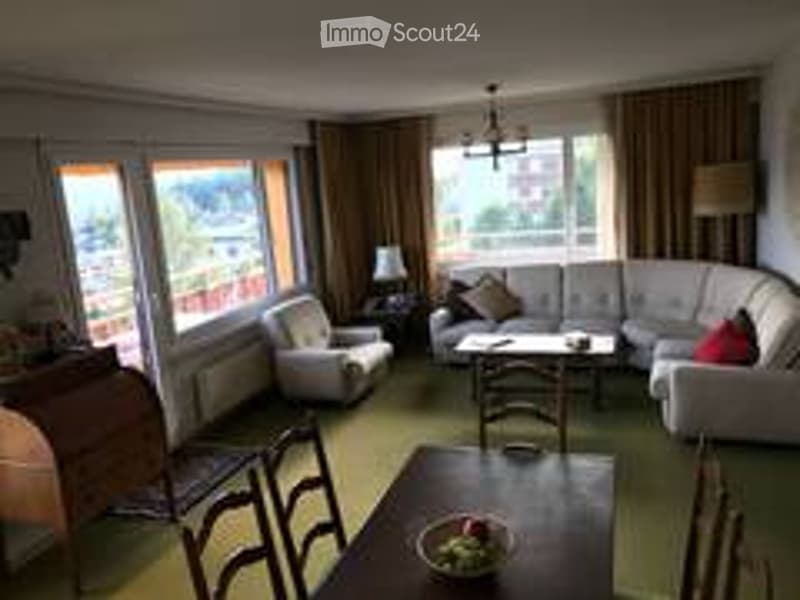 Bright living room with large windows offering a scenic view, furnished with a white sectional sofa, coffee table, armchairs, dining table, and chairs.