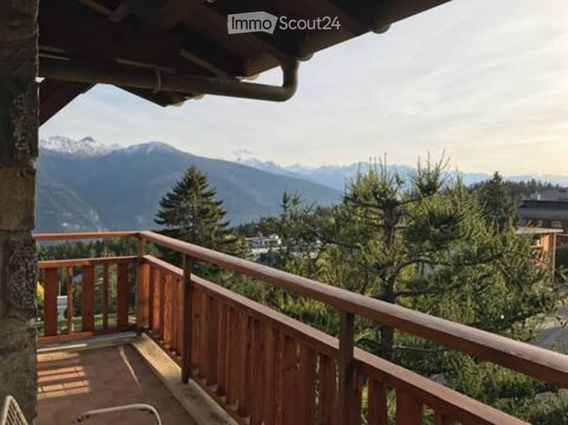 Balcony with wooden railing, overlooking a mountain range and forested area.