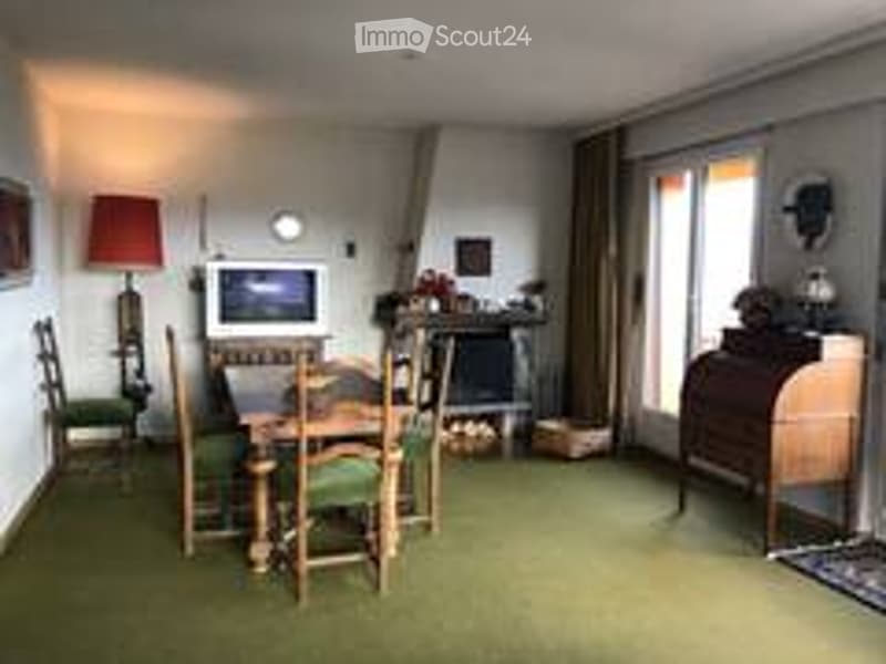 A cozy, well-furnished room with a dining area and a living area. The dining area features a wooden table with four chairs, and the living area includes a sofa and a television. There is a wooden cabinet and a lamp visible, along with large windows covere