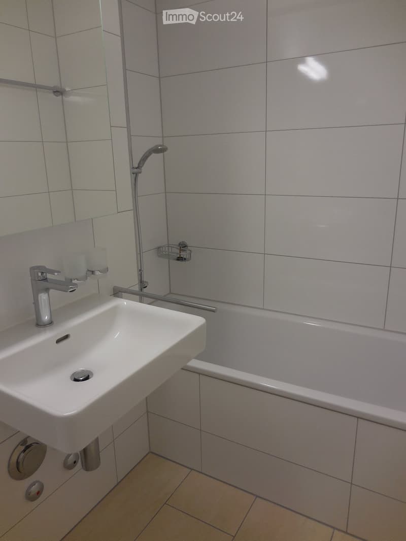 Modern bathroom with a white sink, chrome faucet, bathtub, and showerhead. Light gray tiles on walls and a beige tiled floor.