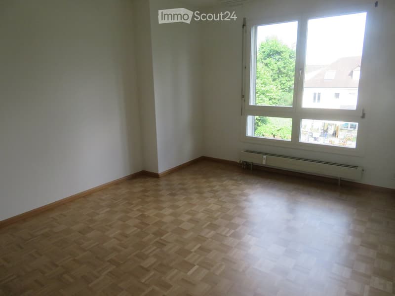 Empty room with wooden parquet flooring, white walls, and large windows offering a view of greenery and neighboring buildings.