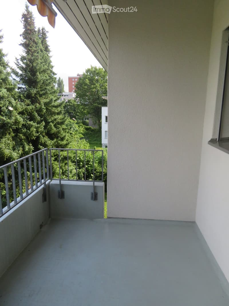 Balcony with metal railing, grey floor, and view of greenery and buildings.