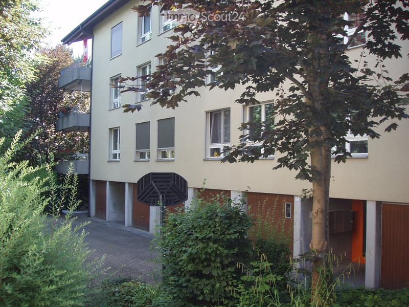 Two-story residential building with light yellow facade, multiple windows, and a small balcony on the upper floor. The building is surrounded by greenery, including trees and shrubs, with a paved driveway leading to a garage entrance.