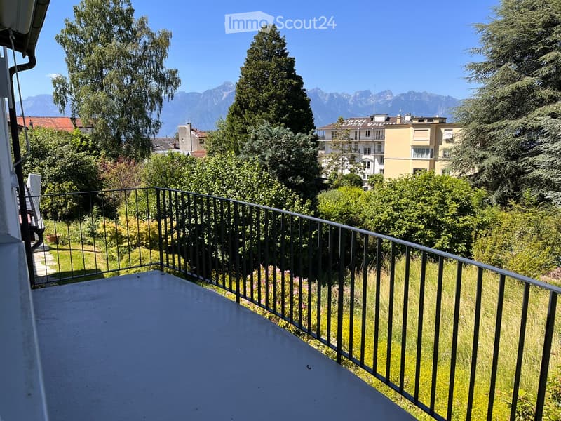 Balcony with metal railing overlooking a green area with trees and distant mountains.