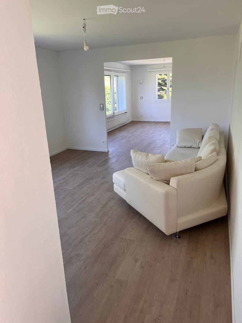 Living room with light-colored wooden flooring, white walls, and a cream-colored sofa with multiple cushions. Large windows allow natural light to enter, and the space is open and minimalistic.