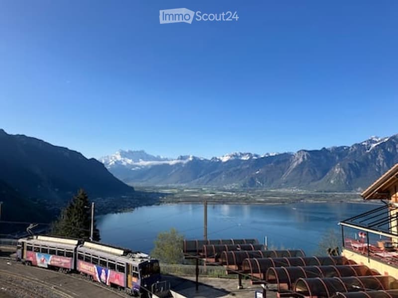 Scenic outdoor view of a lake surrounded by mountains, with a train passing by and a balcony overlooking the landscape.