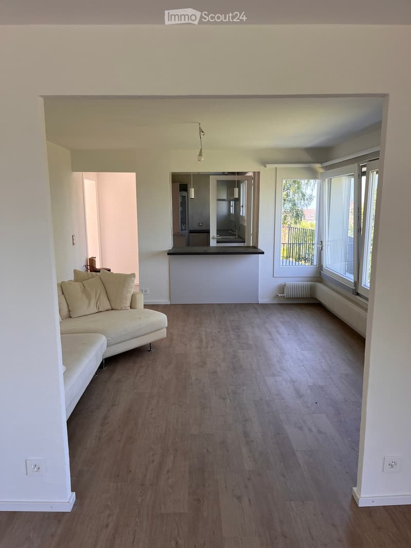 Living room with light-colored sofa, wooden floor, large windows, and a small kitchen area visible in the background.