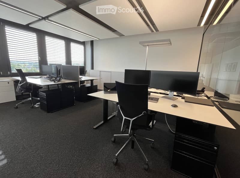 Modern office space with desks, chairs, computers, and office equipment. Large windows provide natural light.
