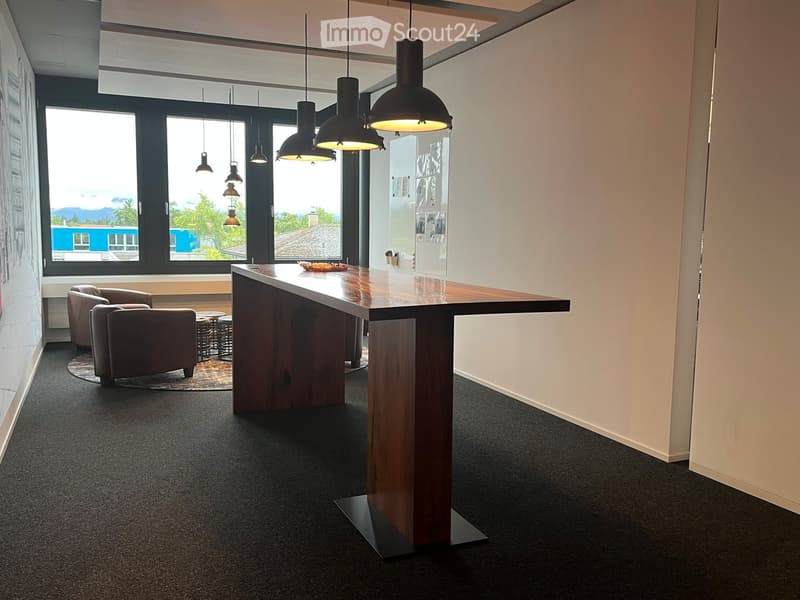 Modern, well-lit room with a long wooden table, two chairs, a small seating area with two armchairs and two stools, and several pendant lights hanging from the ceiling. Large windows offer a scenic view of the outdoors.