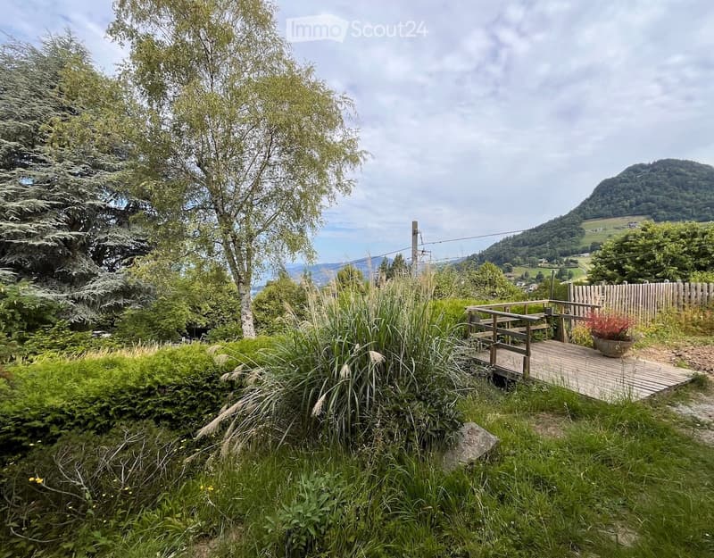 Wooden deck with a bench and potted plant, surrounded by lush greenery and trees, overlooking a scenic valley and hills.