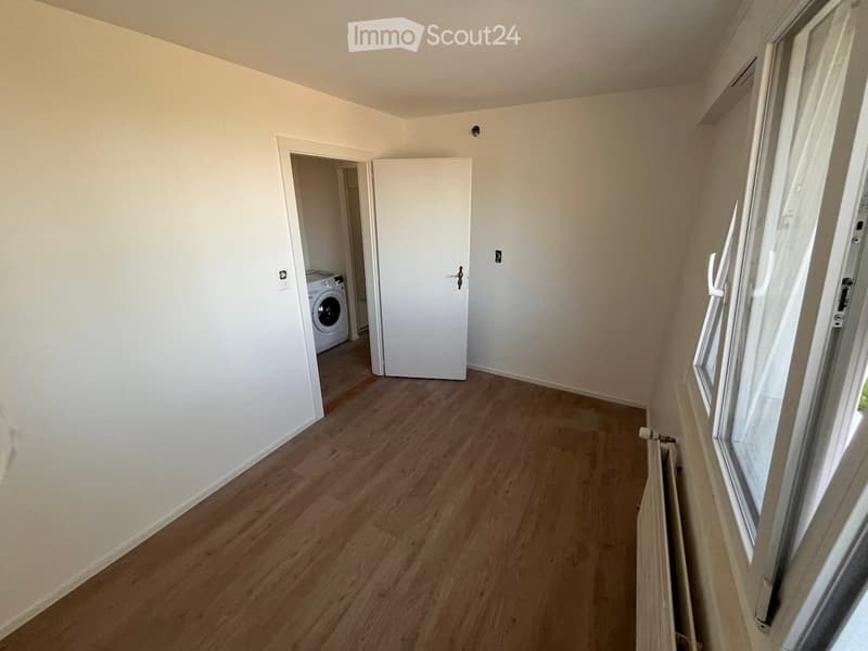 Empty room with light wooden flooring, white walls, and a door leading to a laundry area with a washing machine. A radiator is visible near the window.