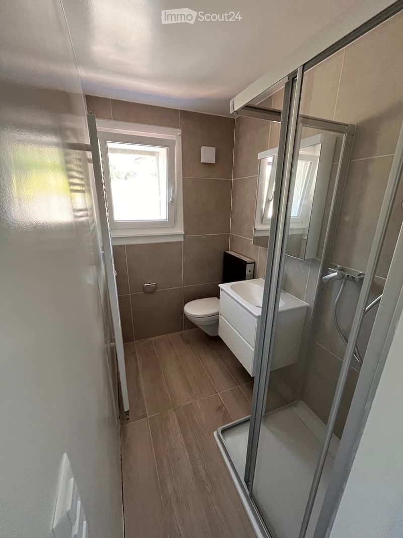 Modern bathroom with wooden flooring, tiled walls, a toilet, a bathtub, a shower, and a window providing natural light.