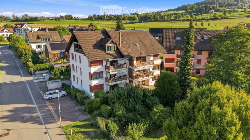 Aerial view of a residential area with multiple houses, balconies, and a parked car. The houses have red-tiled roofs and are surrounded by greenery and open fields.