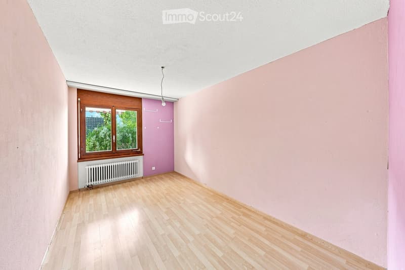 Empty room with light pink walls, wooden flooring, and a window with wooden frame. There is a radiator below the window and a door leading to another room.