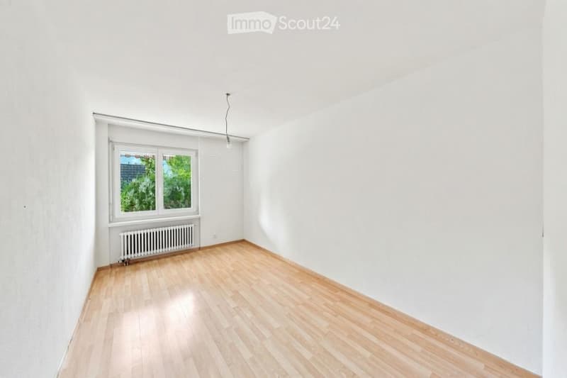 Empty room with light wooden flooring, white walls, and a single window with a radiator below it.