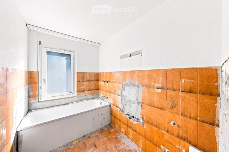 Bathroom with a white bathtub, orange tiles, and a window with frosted glass. The tiles show signs of wear and discoloration.