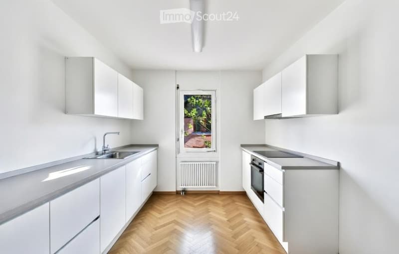 Modern, minimalist kitchen with white cabinets, grey countertops, and a wooden herringbone floor. Features a sink, faucet, and oven.