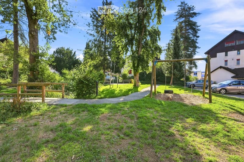 A well-maintained garden area with a swing set, wooden fence, and a paved pathway. The area is surrounded by trees and includes a bench and a few parked cars in the background.