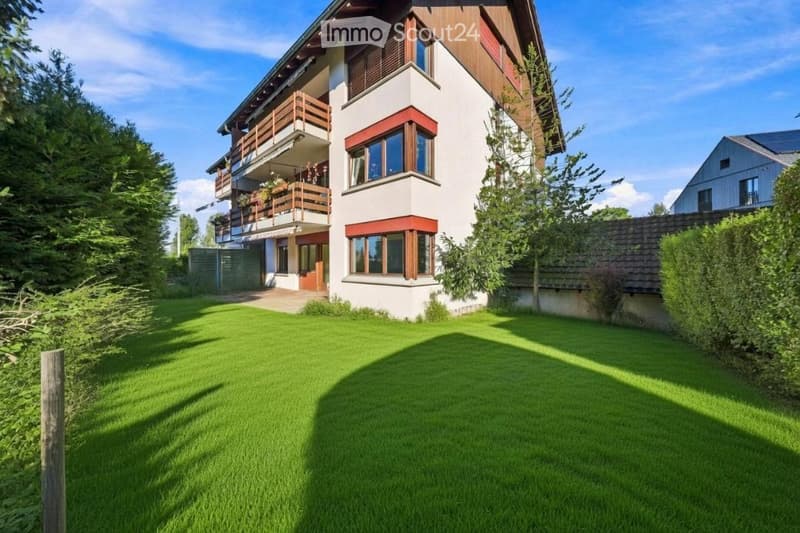 A two-story house with white walls, wooden accents, and large windows. Features include a balcony on the upper floor, a well-maintained lawn, and surrounding trees.