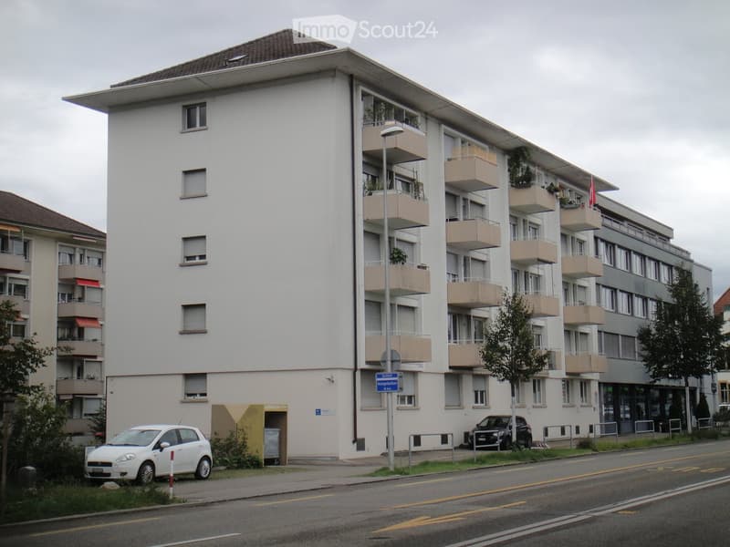 Four-story residential building with multiple balconies, white facade, and beige railings. Two cars are parked in front of the building.