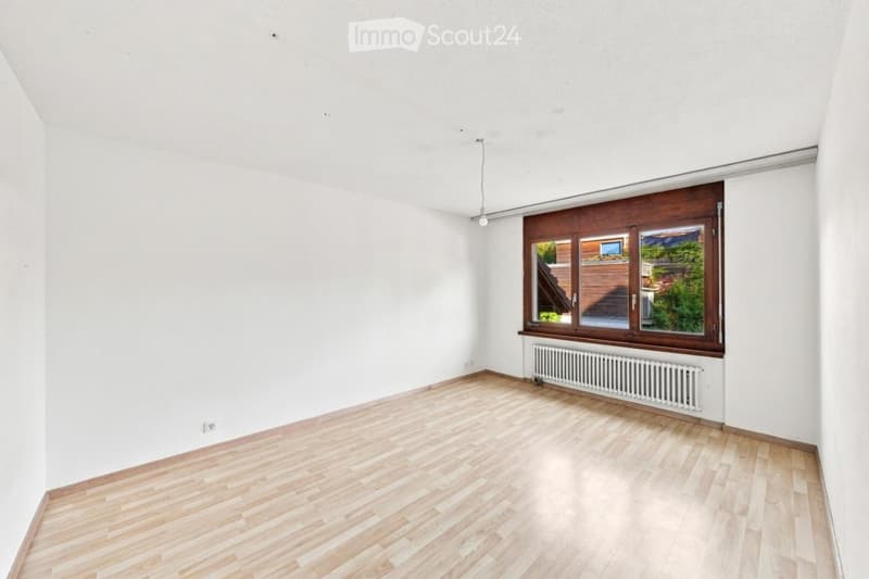 Empty room with light wooden flooring, white walls, and a large window with wooden frame. A radiator is visible below the window.