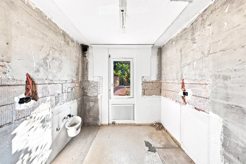Empty room with unfinished walls, a white door, a window showing greenery outside, a radiator, and a white bucket on the floor.