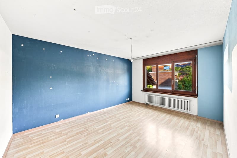 Empty room with wooden flooring, blue walls, and a large window with wooden frame. There is a radiator below the window and visible electrical outlets on the walls.