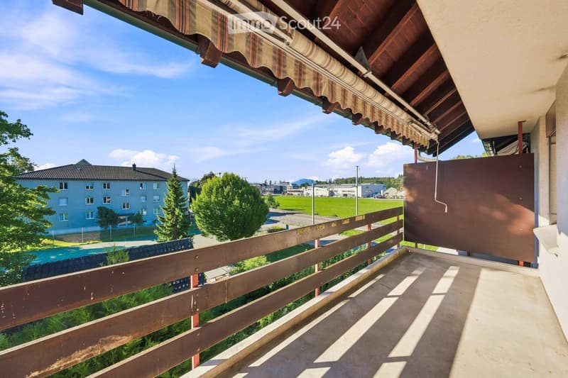 Balcony with wooden railing, striped awning, and scenic view of a green field, trees, and distant buildings.
