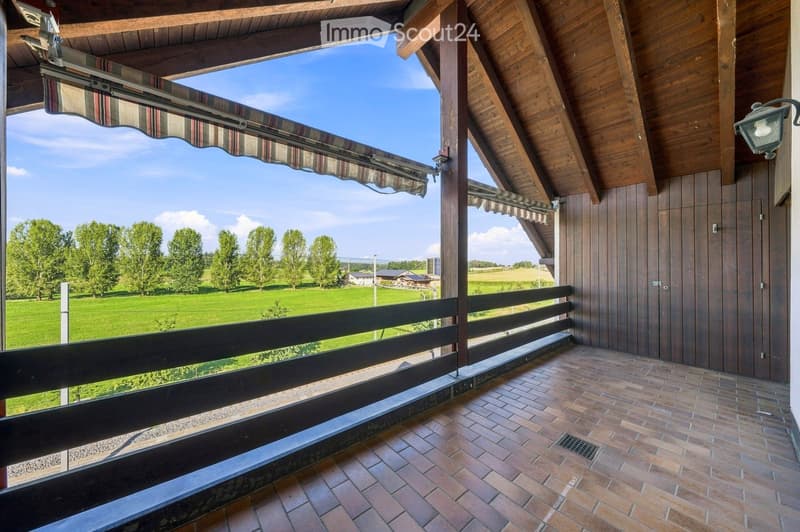 Balcony with wooden beams, brick tiles, and a view of a green field and trees.