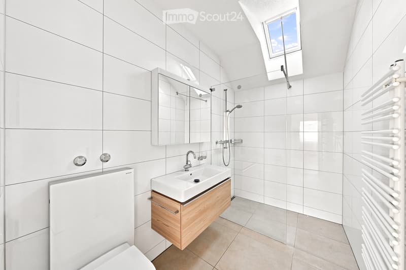 Modern bathroom with white tiles, a wooden vanity, a sink, a mirror, a radiator, and a toilet. Features a skylight window and a glass-enclosed shower area.