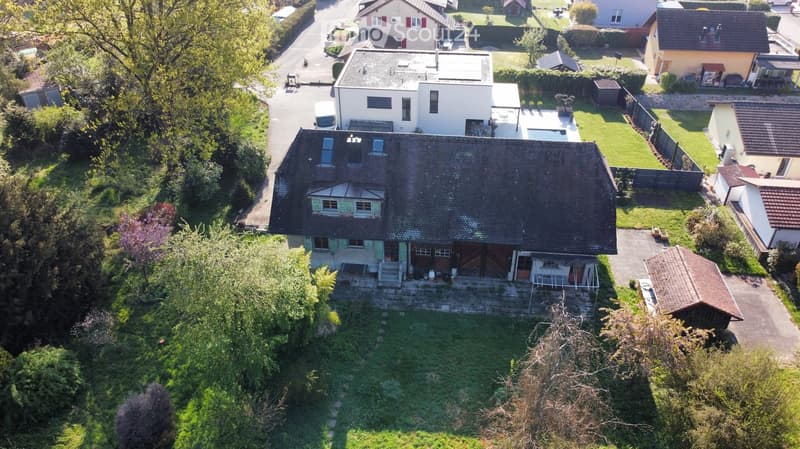 Aerial view of a two-story house with a dark roof and light-colored walls, surrounded by a well-maintained garden and neighboring houses.