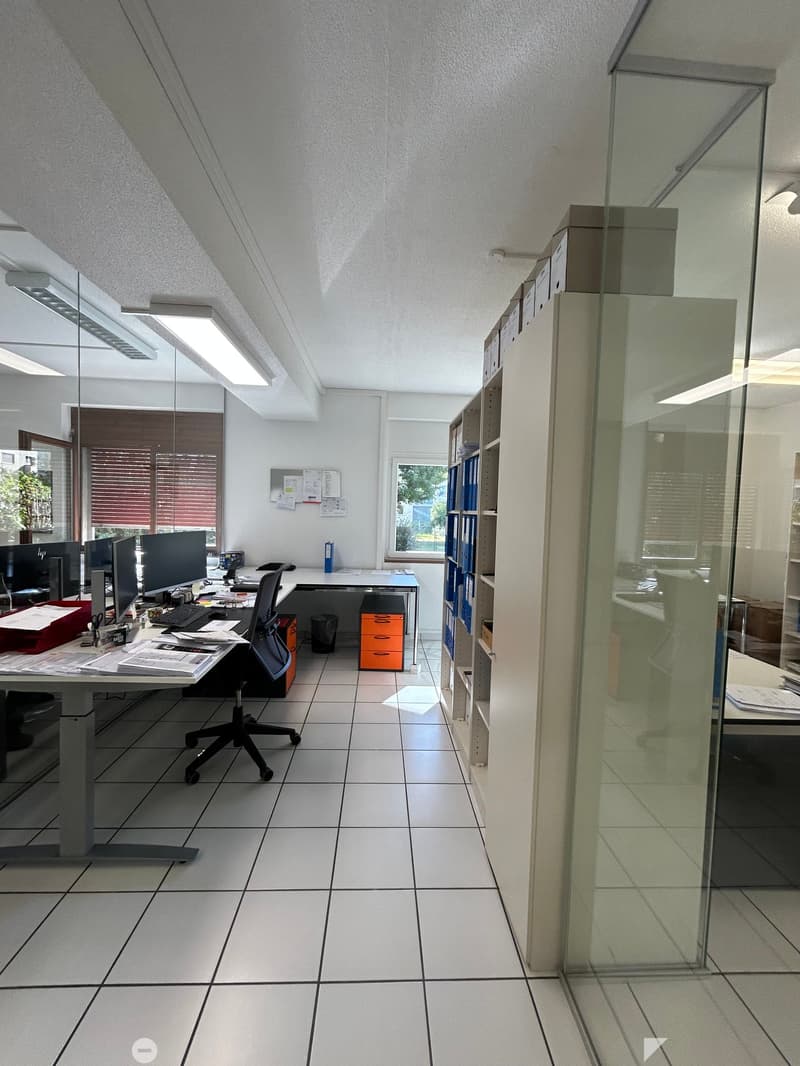 Office space with a desk, chair, computer setup, filing cabinet, and shelves. The room has white tiled flooring and large windows with blinds.