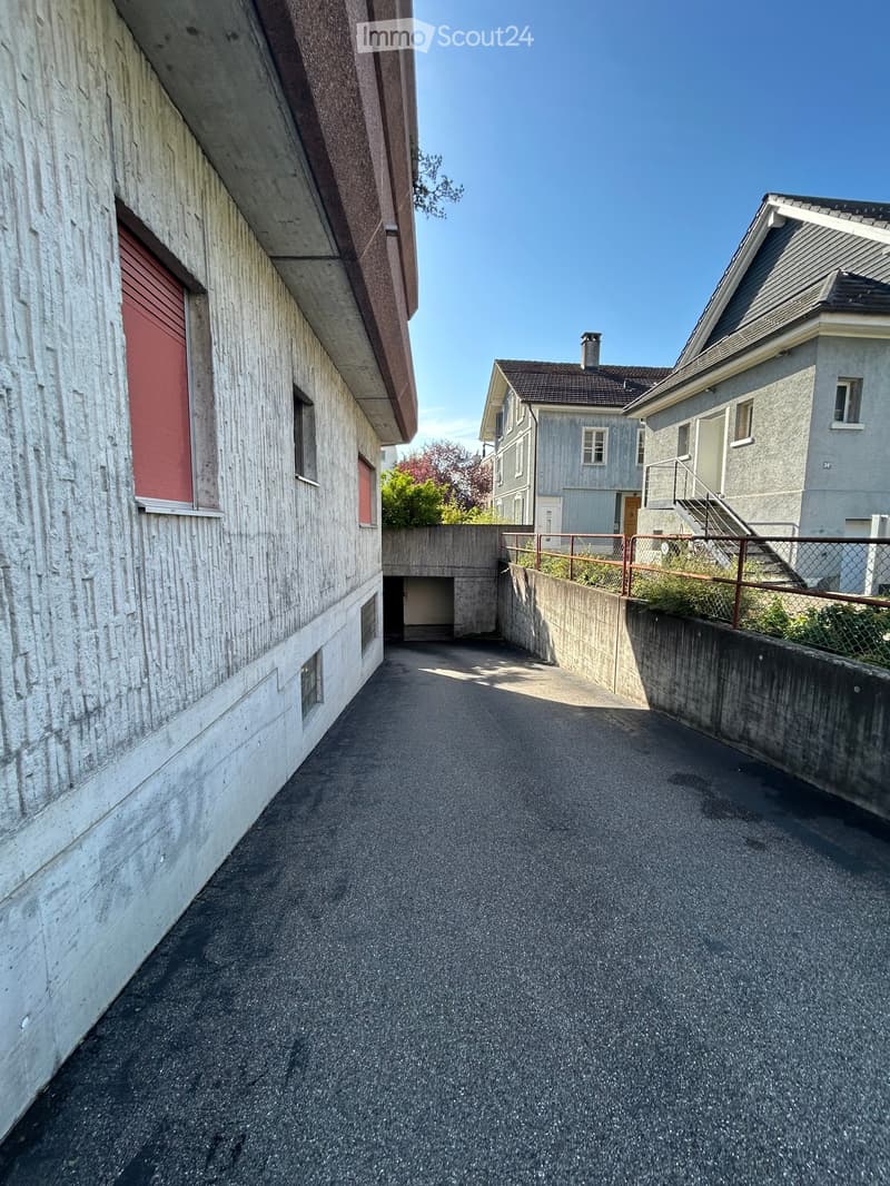 A narrow, paved pathway between two buildings, with concrete walls and a metal fence on one side.