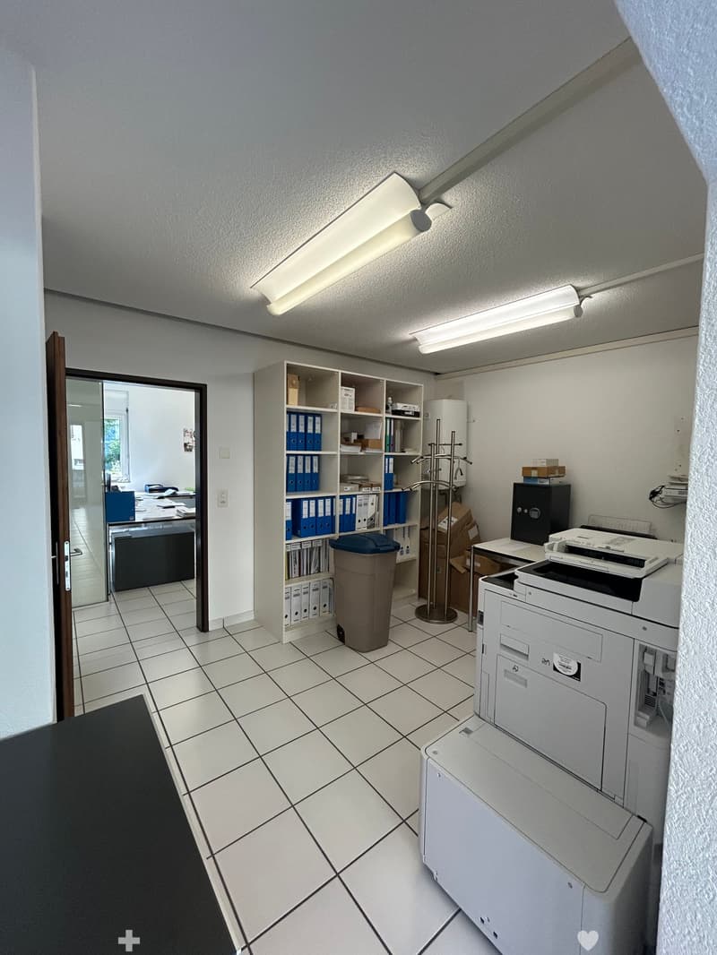 Office space with tiled floor, white walls, fluorescent lighting, shelves with binders, printer, copier, and trash bin.