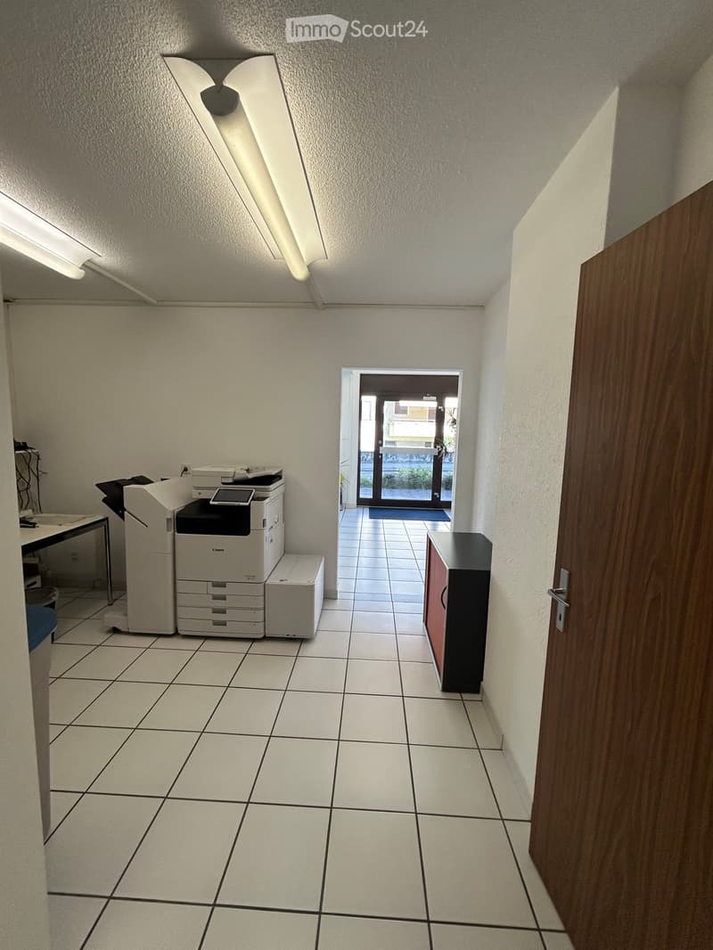 Office area with tiled floor, printer, table, and cabinet. An open doorway leads to another room with a tiled floor and a small table.