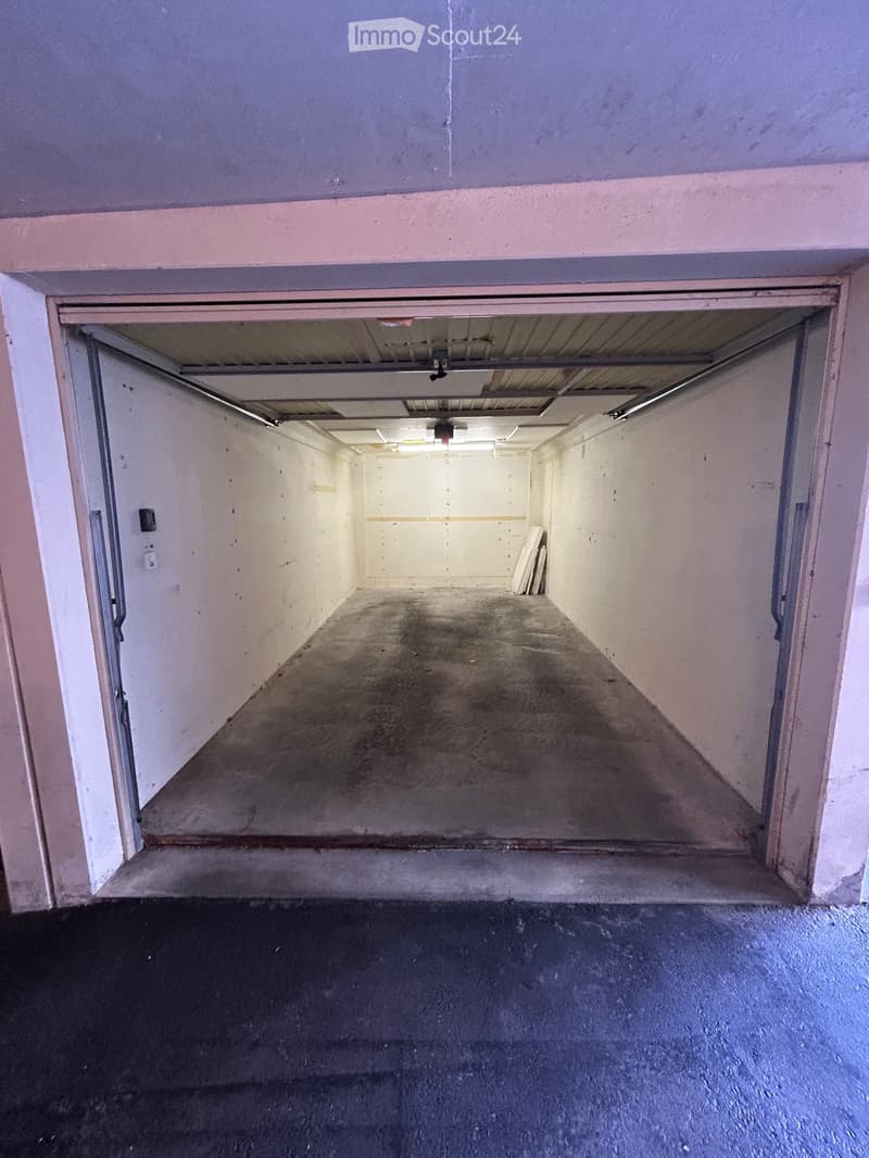 Empty garage space with concrete floor, white walls, and a partially open garage door leading to an outdoor area.