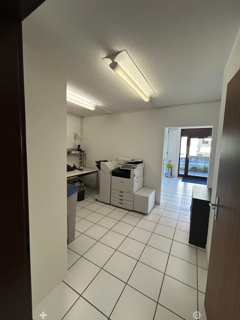 Office space with tiled floor, white walls, fluorescent lighting, printer, table, and chair.