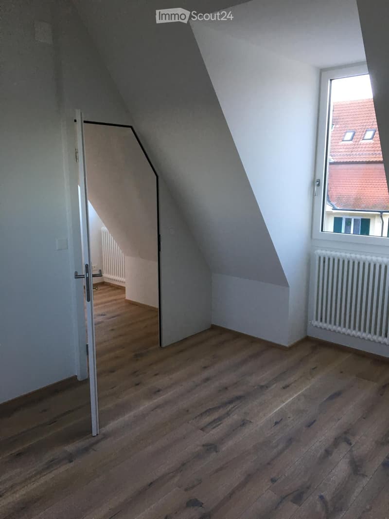 Empty attic room with wooden flooring, white walls, and a window showing a neighboring building with a red roof.