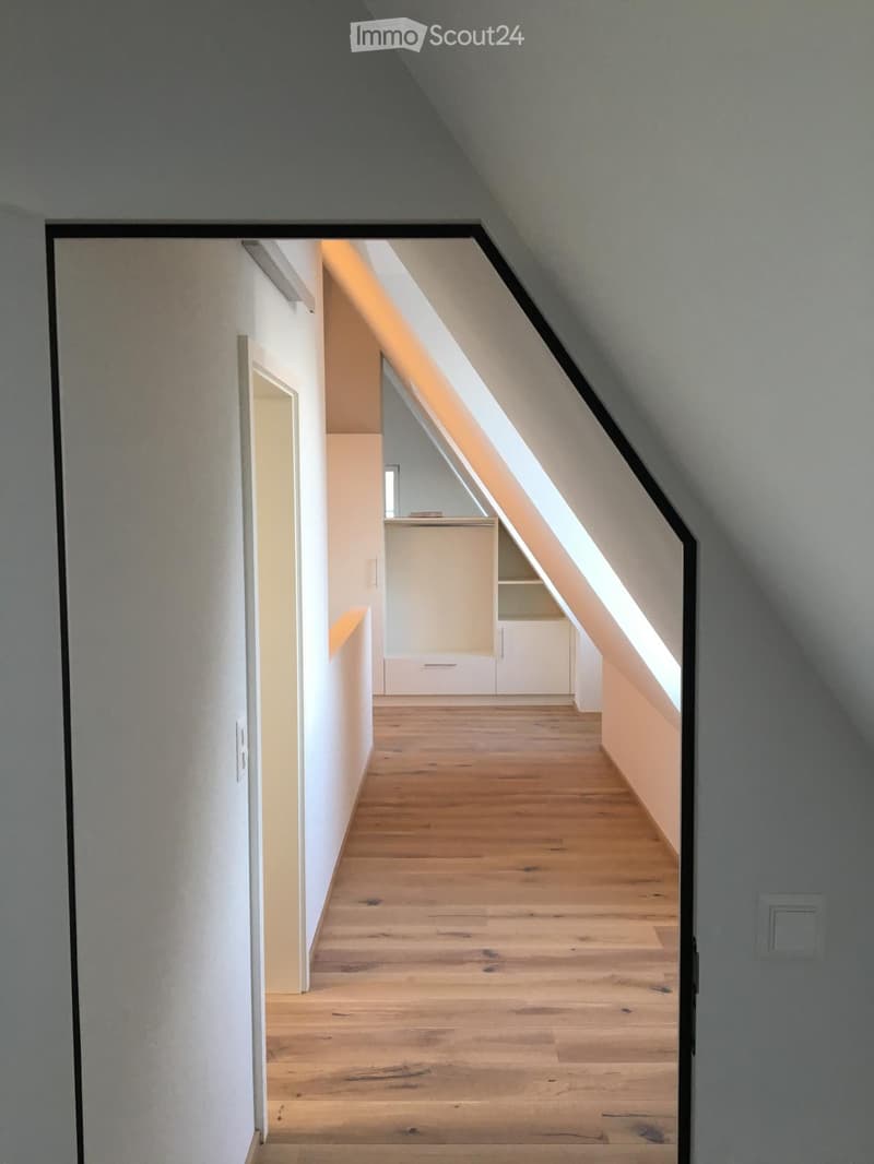 A narrow, well-lit hallway with wooden flooring and white walls, leading to a small closet area with built-in storage under a sloped ceiling.