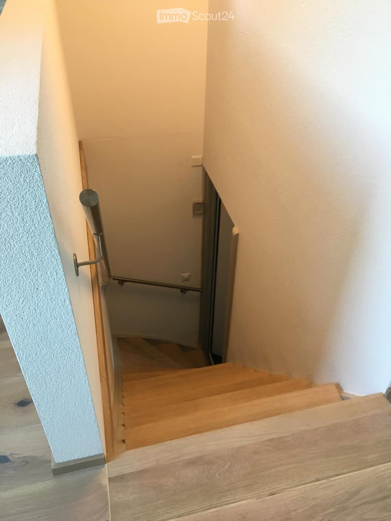 Wooden staircase with a handrail, leading upwards, with light-colored walls and wooden flooring.
