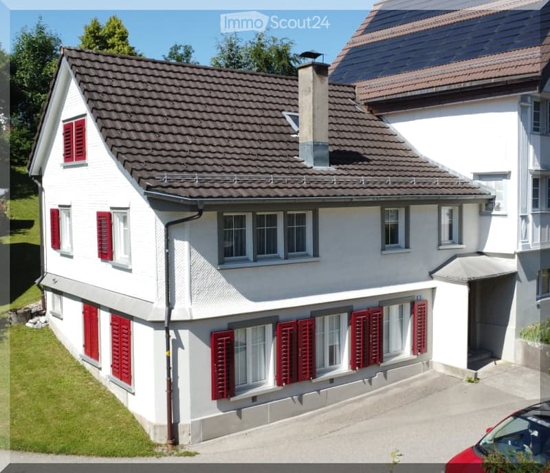 Two-story white house with red shutters, multiple windows, and a chimney. The house has a tiled roof and is surrounded by greenery.