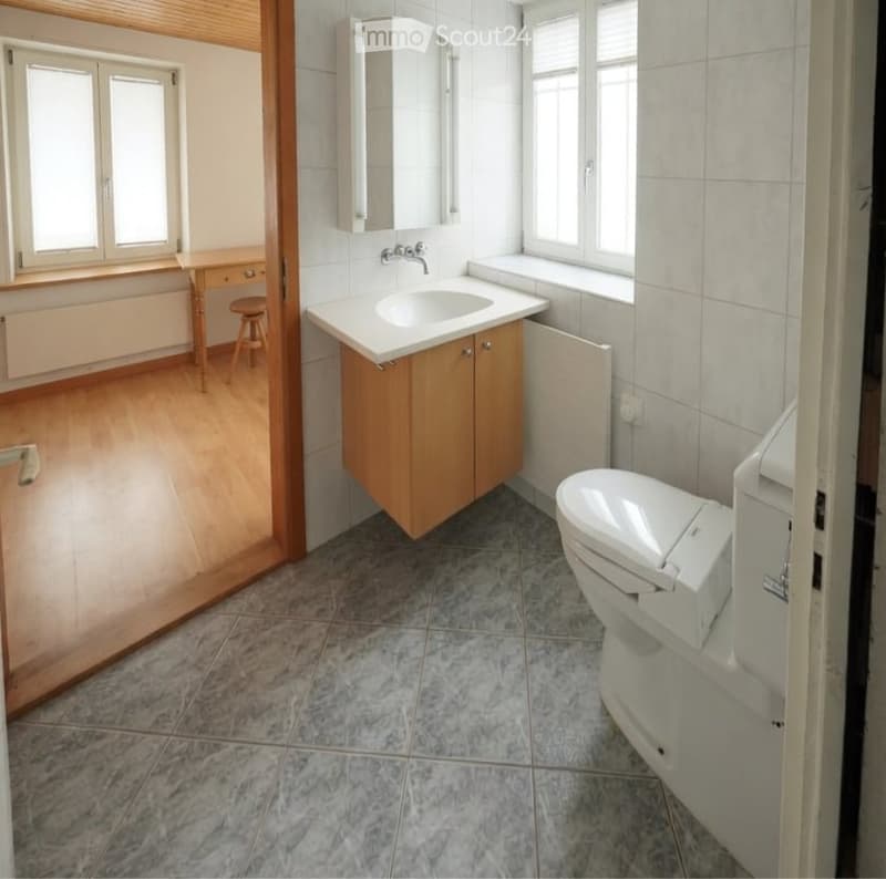 Bathroom with a sink, toilet, mirror, wooden cabinet, and a small table with a stool in the adjacent room.