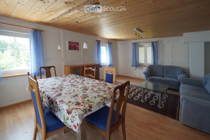 Bright living and dining area with wooden floors, exposed wooden ceiling beams, large windows with blue curtains, a blue sofa, a glass coffee table, a dining table with floral tablecloth and chairs, and a wooden cabinet.