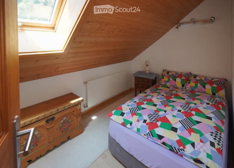 Cozy attic bedroom with wooden slanted ceilings, skylight window, colorful patchwork quilt on the bed, wooden nightstand with lamp, and a decorative wooden chest.