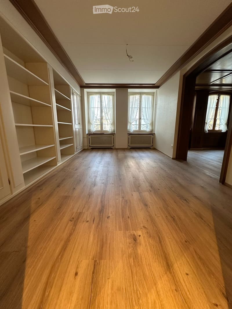 Empty room with wooden flooring, built-in shelves on one wall, and large windows with curtains. Radiators are visible beneath the windows.