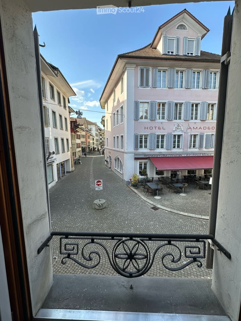 Balcony overlooking a narrow cobblestone street lined with buildings, including a hotel and restaurant with outdoor seating.