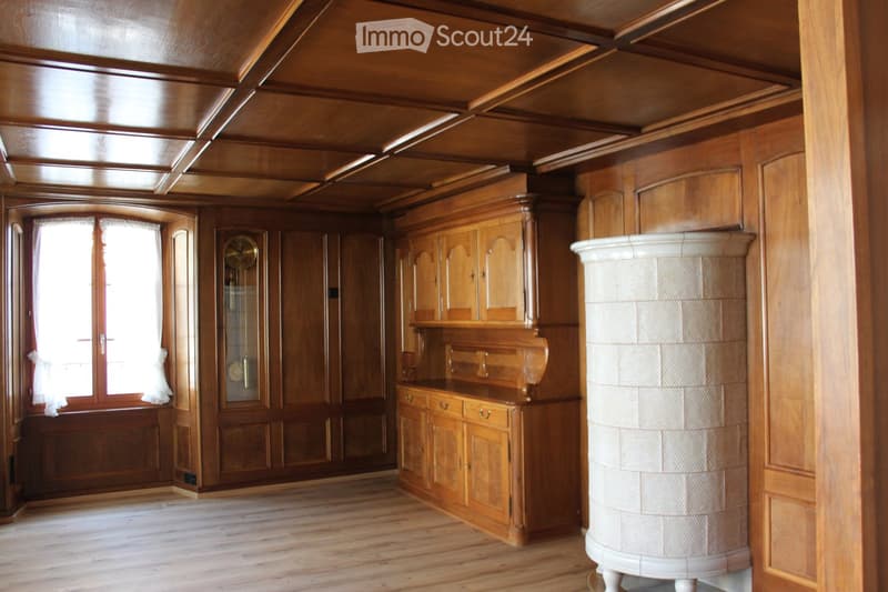 An unfurnished room with wooden walls, ceiling, and flooring, featuring built-in wooden cabinets, a large window with white curtains, and a decorative white column.