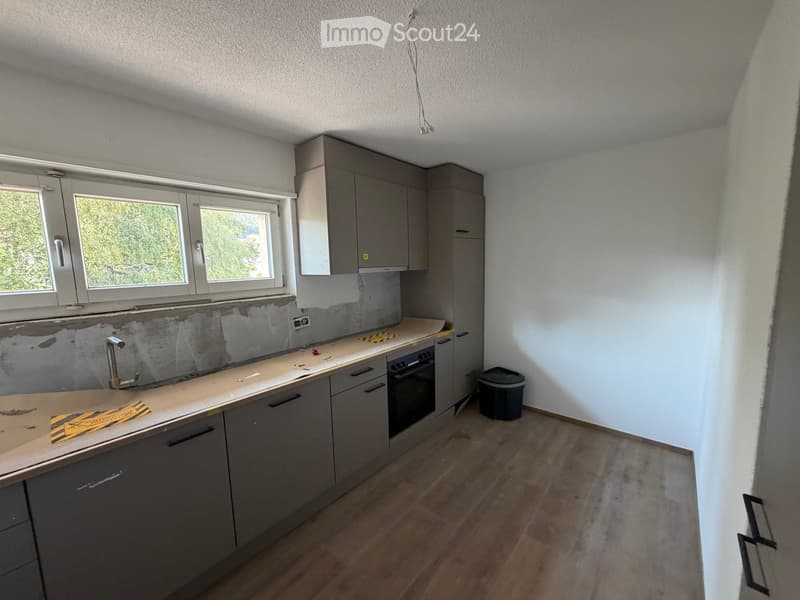 Empty kitchen with grey cabinets, a countertop, and a sink. The backsplash is partially removed, revealing the wall underneath.